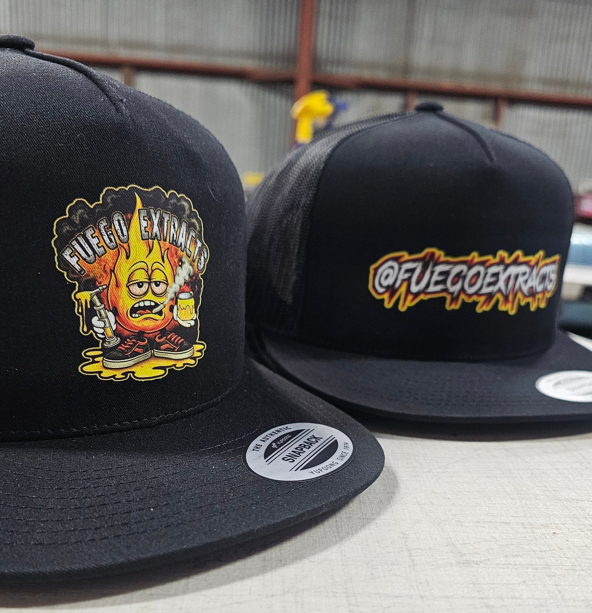 Two black flat-brim snapback caps on a light floor in a warehouse-like setting. The cap in front has a colorful cartoon flame graphic with the text 'Fuego Extravagante' on the front.