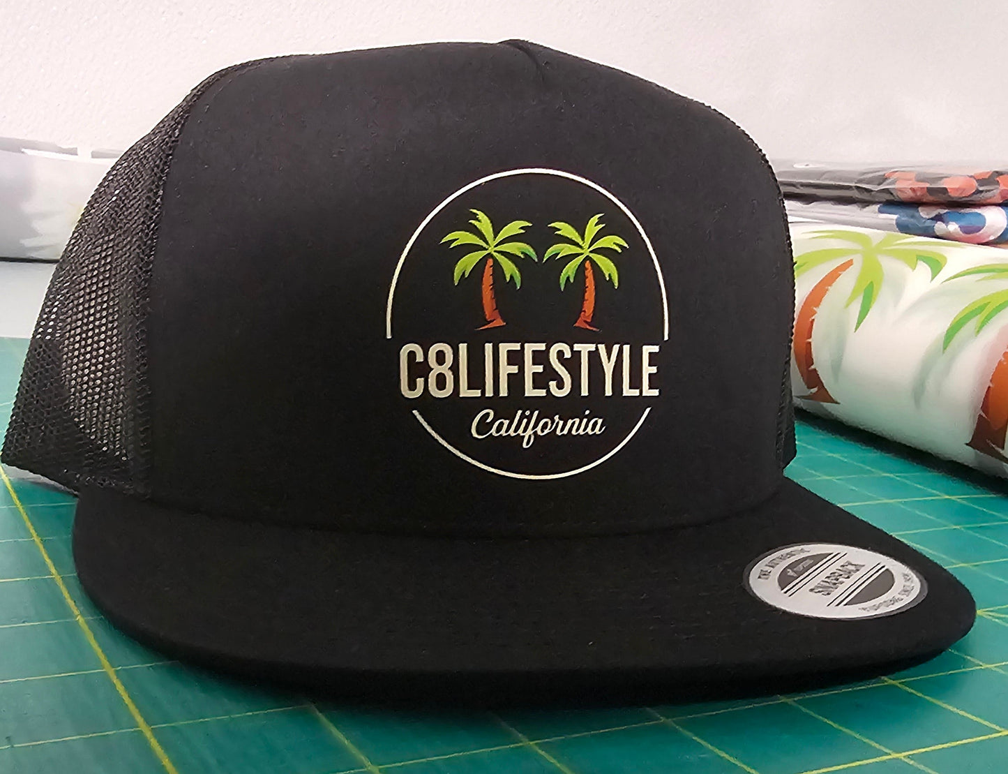 A black trucker hat with a circular 'C8LIFESTYLE California' palm tree logo on the front, resting on a green cutting mat.