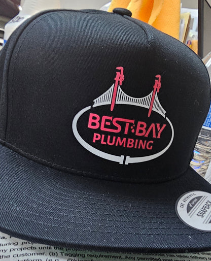 A black snapback cap featuring a circular logo on the front that reads 'BEST BAY PLUMBING' in red and white, with a bridge graphic above. The cap is photographed in a cluttered indoor setting with papers, a yellow cloth, and household items visible in the background.